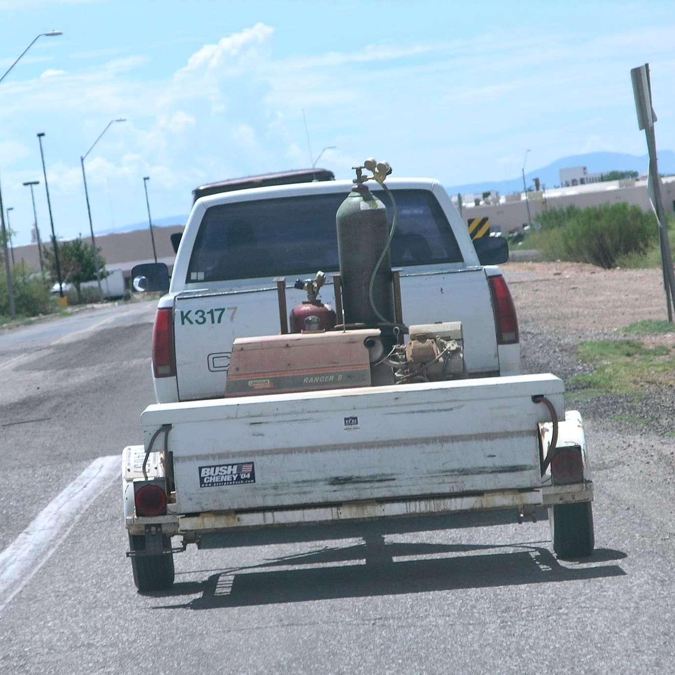 A white pick up with the identifier K3177 pulling a trailer with welding equipment and a Bush-Cheney 2004 campaign stick (a Hatch Act violation).