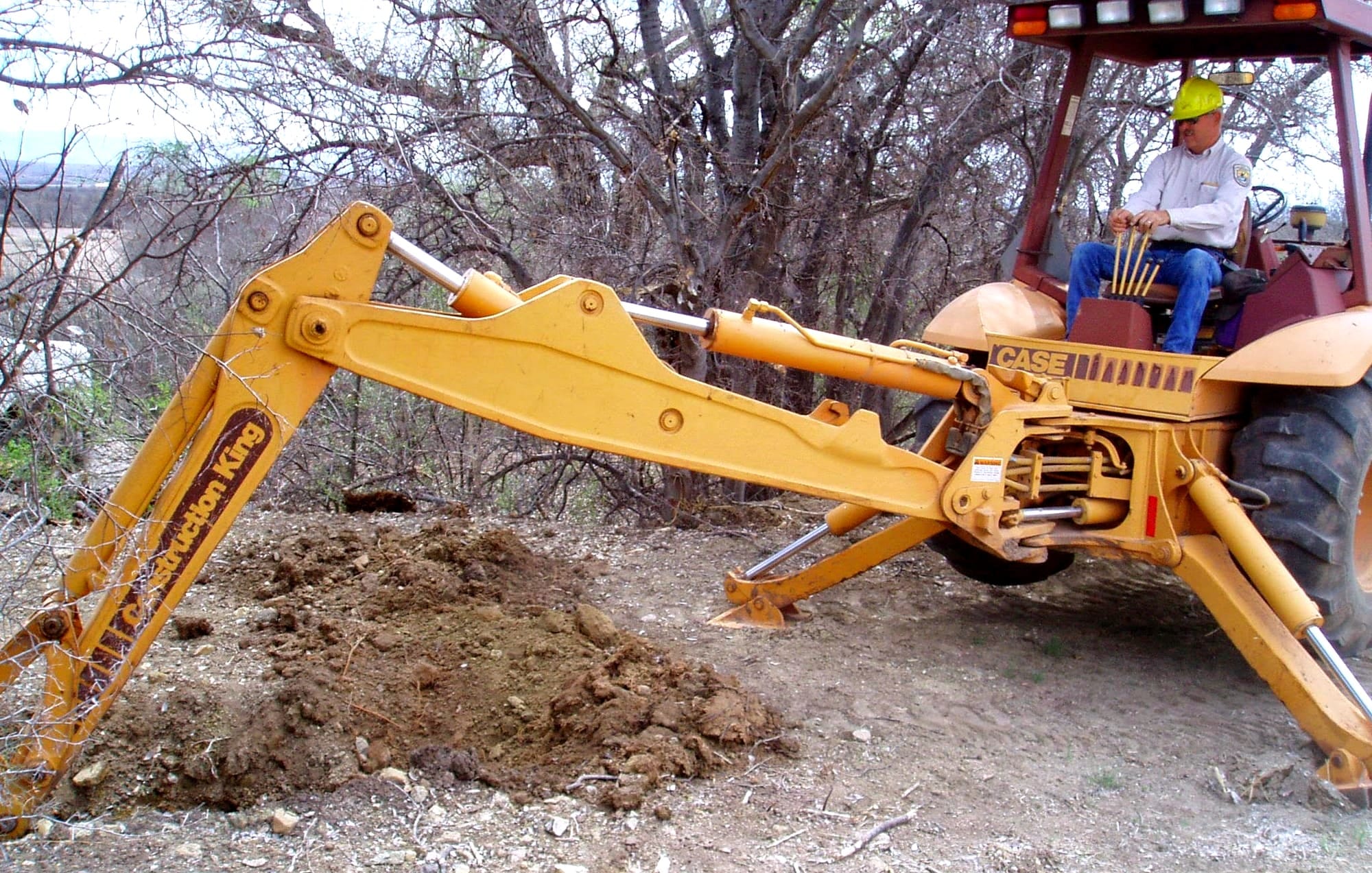 A man sits on a backhoe digging into dirt, with short trees in the background.
