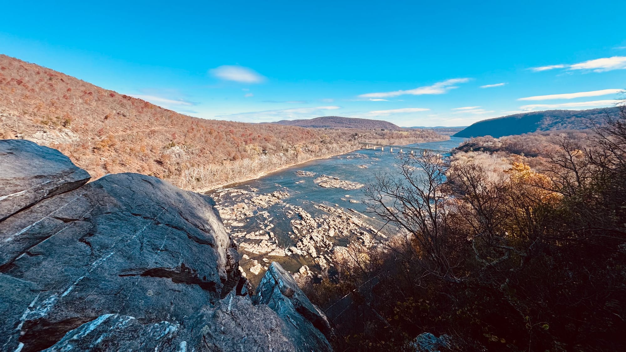View from a rocky outcrop overlooking a rocky river below, with wooded mountains around, and mostly clear blue skies with a few distant clouds.