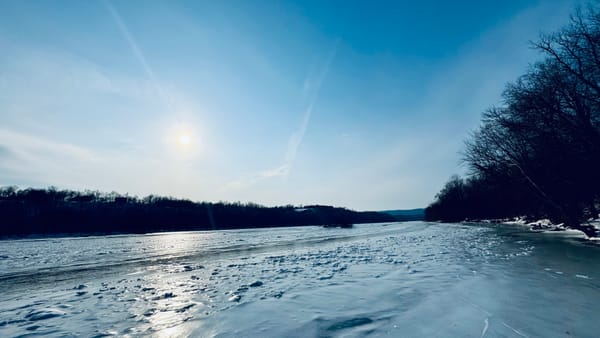A frozen-over river is lined by trees, with partly cloudy skies overhead.